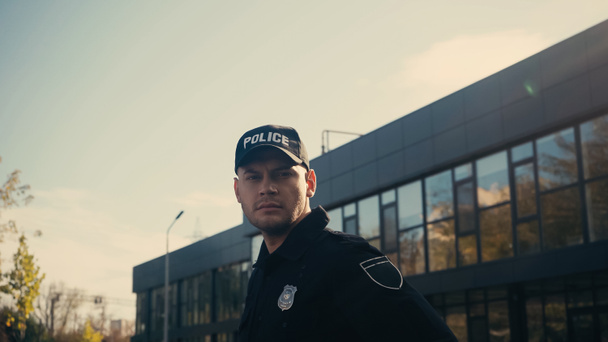 policial sério de pé em boné e uniforme enquanto olha para a câmera perto do edifício   - Foto, Imagem