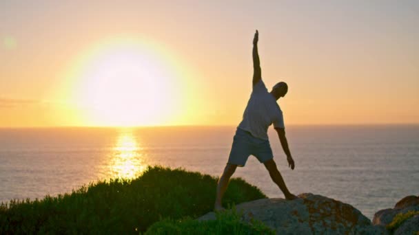 Izmos atléta sziluett nyúlik a tenger felkelő nap. Erős ember gyakorol a sziklaóceán szélén. Ismeretlen jógás fickó, aki egyedül élvezi a naplementét. Aktív sportéletmód koncepció. - Felvétel, videó