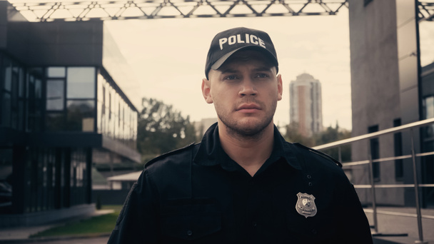 серьезный полицейский в кепке и униформе патрулирующий современную улицу - Фото, изображение