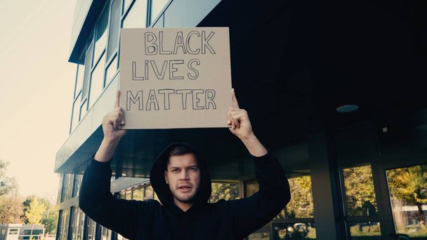 serious young activist in hoodie holding placard with black lives matter lettering on street - Фото, изображение