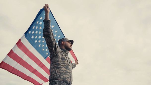 Vista de ángulo bajo de soldado en uniforme y gorra sosteniendo bandera americana al aire libre  - Foto, imagen