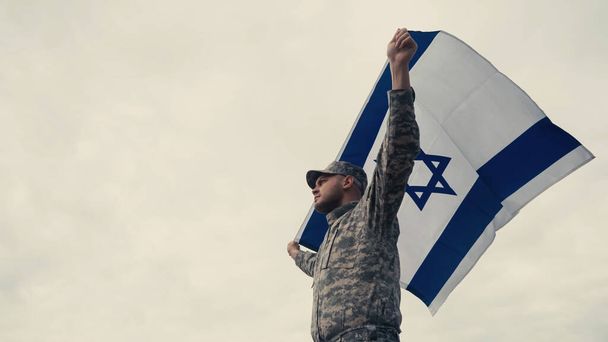 Low angle view of soldier in uniform holding national flag of Israel with sky at background  - Fotó, kép