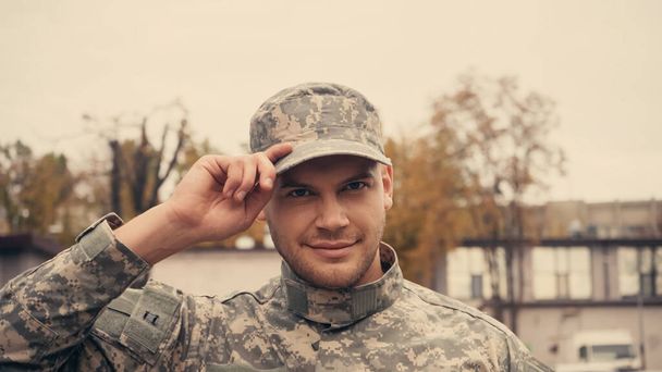 Soldado sorridente em uniforme ajustando tampa e olhando para a câmera ao ar livre  - Foto, Imagem