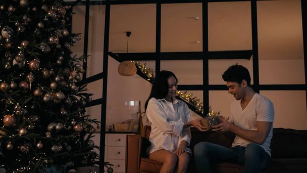 happy woman presenting gift to cheerful boyfriend near christmas tree   - 写真・画像