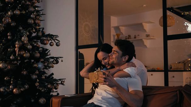 happy woman hugging boyfriend with present sitting on couch near christmas tree - Fotó, kép