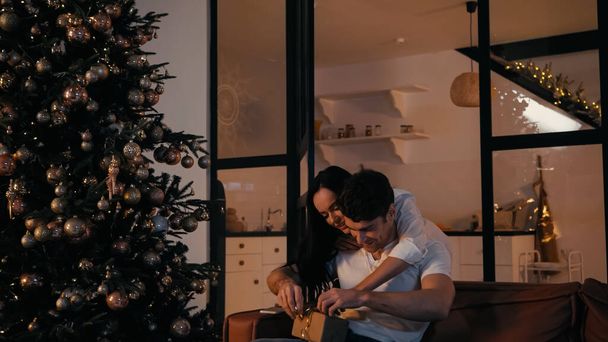 happy woman helping to open present of boyfriend near christmas tree  - 写真・画像