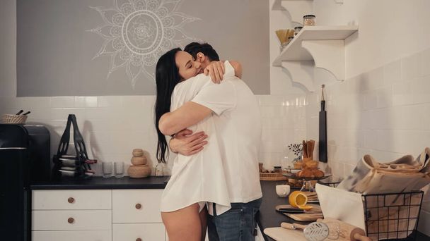 brunette young woman in white shirt smiling while embracing boyfriend in modern kitchen - Foto, imagen