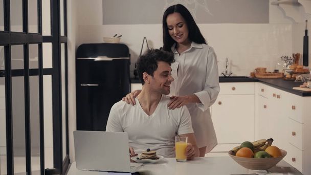 mujer sexy abrazando hombros de hombre feliz cerca del desayuno y el ordenador portátil en la cocina - Foto, Imagen