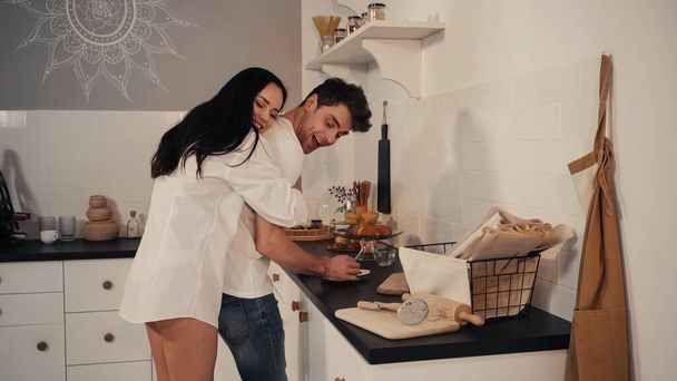 happy young woman in white shirt hugging cheerful boyfriend in kitchen  - Zdjęcie, obraz