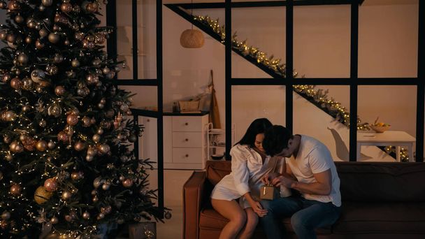 happy man opening present near joyful girlfriend sitting on couch near christmas tree  - Valokuva, kuva