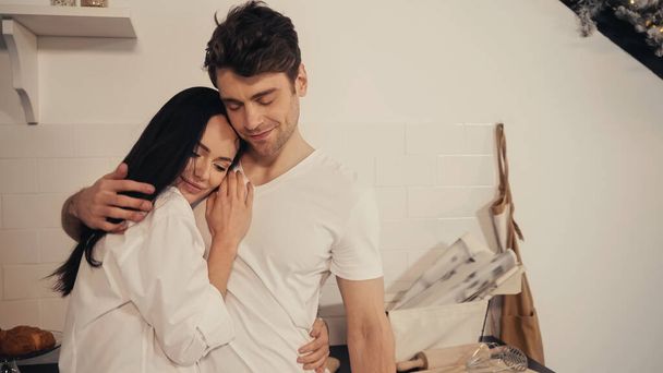 brunetta giovane donna in camicia bianca abbracciando fidanzato soddisfatto in cucina moderna  - Foto, immagini