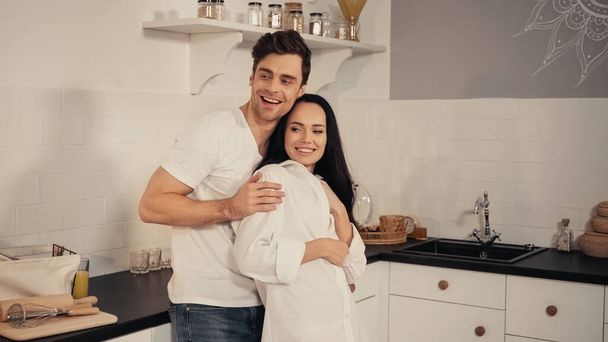 szczęśliwy człowiek obejmujący wesoły dziewczyna i uśmiechnięty w kuchni - Zdjęcie, obraz