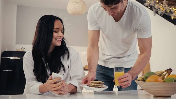 man serving pancakes and orange juice to happy girlfriend in kitchen with christmas decoration  - Foto, Imagen