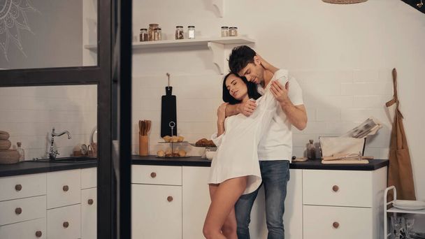 男抱擁ブルネット若いです女性で白いシャツで現代的なキッチンに立っている間   - 写真・画像