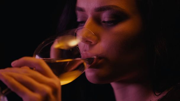 Молодая женщина пьет белое вино из стакана, изолированного на черном  - Фото, изображение