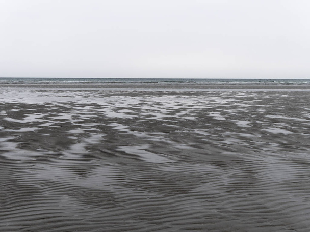 Υγρή κυματιστή άμμος στην ακτή σε χαμηλή παλίρροια σε συννεφιασμένη μέρα. Θλιμμένο παραθαλάσσιο τοπίο. Η σκληρή φύση της Βόρειας Ευρώπης. Η κρύα ακτή του Ατλαντικού Ωκεανού. - Φωτογραφία, εικόνα