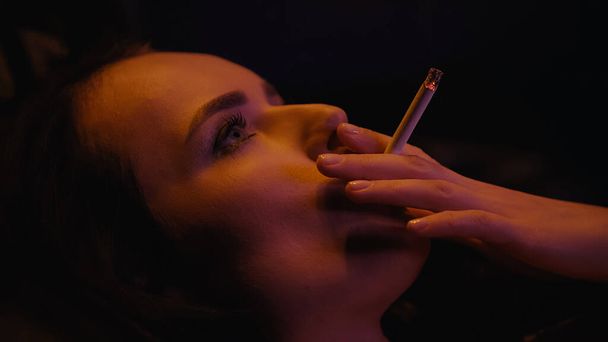 Νεαρή γυναίκα κοιτάζει αλλού ενώ καπνίζει, απομονωμένη στο μαύρο.  - Φωτογραφία, εικόνα