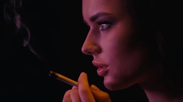 Vista lateral de una mujer joven sosteniendo un cigarrillo borroso sobre un fondo negro  - Foto, Imagen