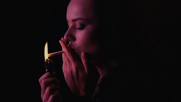 Junge Frau hält Feuerzeug und Zigarette isoliert auf schwarz mit Beleuchtung  - Foto, Bild