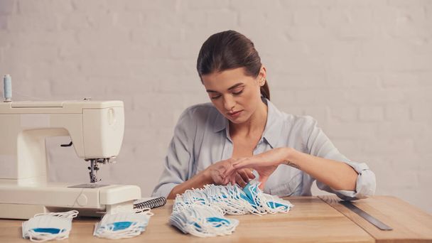 Morena costurera contar máscaras médicas cerca de la máquina de coser y regla en atelier  - Foto, Imagen