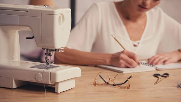 Vista recortada de la máquina de coser cerca de anteojos y costurera borrosa en atelier  - Foto, imagen