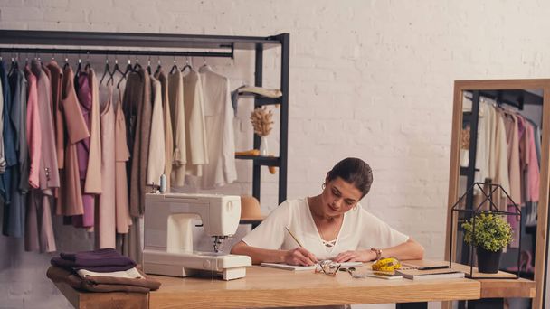 Designer writing on notebook near cloth and sewing machine in atelier  - Φωτογραφία, εικόνα