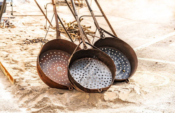 Tres grandes sartenes de metal con agujeros para asar castañas se colocan en la arena - Foto, Imagen