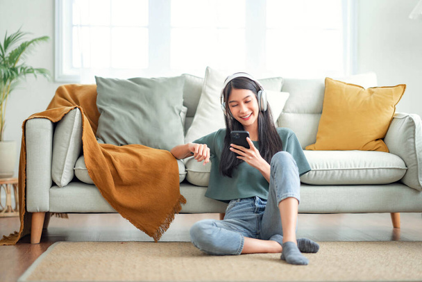 Felice donna asiatica ascoltando musica dal telefono cellulare mentre seduto sul tappeto accanto al divano a casa, Ragazza sorridente rilassante con le cuffie al mattino, Tempo di rilassarsi. - Foto, immagini