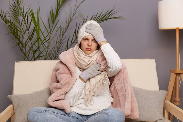 Indoor shot van zieke ongezonde bevroren vrouw dragen pet, handschoenen, sjaal en jas zitten in de woonkamer, valt verschrikkelijke hoofdpijn en griep symptomen, zeer koud in de winter. - Foto, afbeelding
