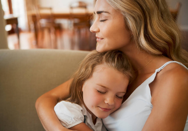 Young loving caucasian mother hugging her little daughter relaxing and sitting on the couch together at home. Innocent carefree girl embracing her mom resting on the couch. - Фото, зображення