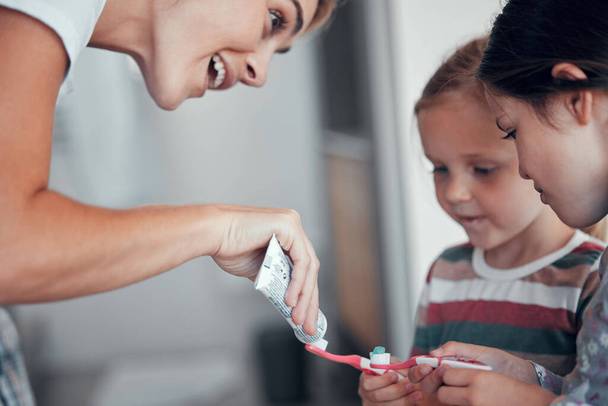 Caucasian mom squeezing toothpaste from a tube onto toothbrushes of two little girls in pyjamas at home. Teaching kids good hygiene habits. Brush twice daily to prevent tooth decay and gum disease. - Photo, Image