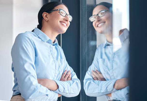 Confident empowered mixed race businesswoman wearing glasses and looking out a window with arms crossed in an office. One female only thinking and dreaming about goals, vision and success for startup. - 写真・画像