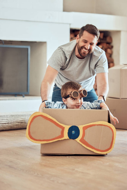 Ευτυχισμένος ανύπαντρος πατέρας σπρώχνει το παιδί του σε χάρτινο κουτί στο σπίτι. Αξιολάτρευτο αγοράκι που κάθεται σε αυτοσχέδιο αεροπλάνο και παίζει με ανύπαντρο γονιό σε ένα σαλόνι στο νέο του σπίτι.. - Φωτογραφία, εικόνα