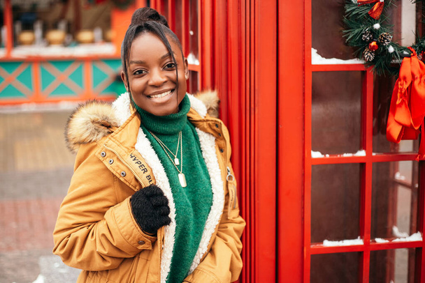 Mujer afroamericana parada calle afuera cerca del mercado justo del árbol de Navidad, cabina telefónica roja. Ambiente de Año Nuevo, vacaciones de invierno, clima frío - Foto, imagen