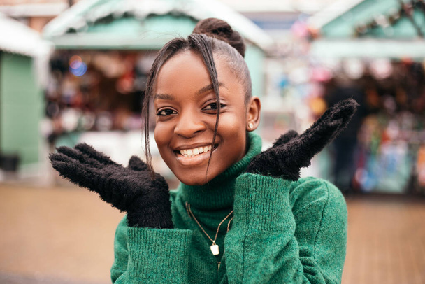 驚いたことに、ショックを受けたアフリカ系アメリカ人女性が公正なクリスマスマーケットの外の通りに立って、笑顔ポーズ。新年の雰囲気、冬の休暇、寒い天候 - 写真・画像