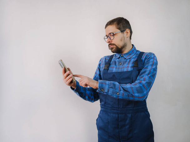 Een jongeman in een blauw werkpak die reparatieappartement doet. Huisrenovatie concept. Man met smartphone sms 'en en afluisteren op mobiele telefoon, op zoek naar oplossing op internet, het bestellen van goederen voor reparaties - Foto, afbeelding