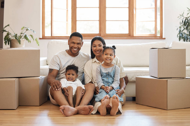 Портрет счастливой молодой семьи с двумя детьми, сидящими в новом доме, сложенные картонные коробки в гостиной. Смешанная раса пары с детьми, покупающих недвижимость, дом или квартиру. - Фото, изображение