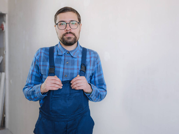 young man in blue work suit looking at camera on white wall background. Repair apartment. Home renovation concept. - Photo, Image