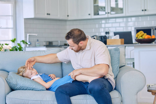 Caucasian father using his hand to feel the high body temperature on his sick little daughters forehead for symptoms of fever, flu or covid. Worried parent caring for ill child lying on sofa at home. - Photo, Image