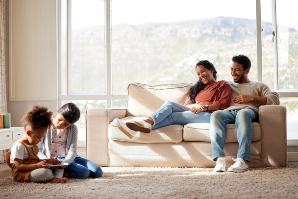 Happy family relaxing at home. Smiling young parents with girls on a sofa, watching their cute little children play and have fun with a digital tablet on the floor in the living room with a window. - Foto, imagen