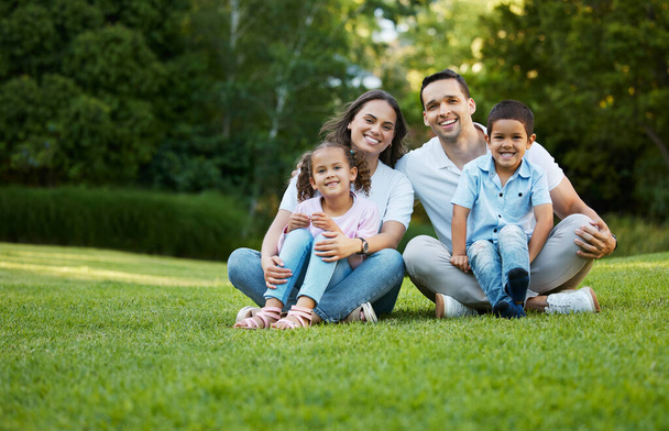 Портрет беззаботной смешанной расовой семьи, проводящей время вместе в парке. Счастливые родители с сыном и дочерью сближаются и веселятся на открытом воздухе. Молодая пара сидит с двумя детьми, сидя на траве. - Фото, изображение