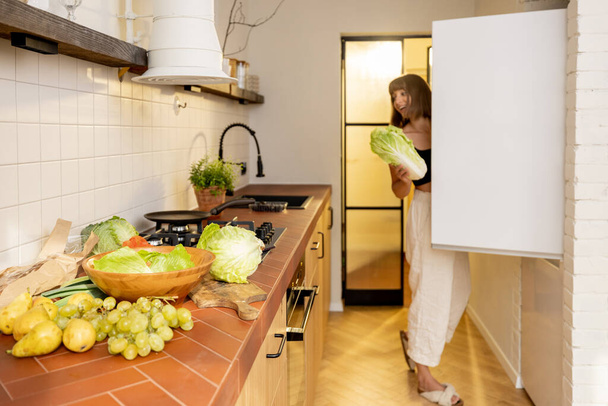 Young woman cooks healthy vegetarian food with green fresh ingredients, looking into a fridge in kitchen at home. Healthy lifestyle and eating concept - Fotó, kép
