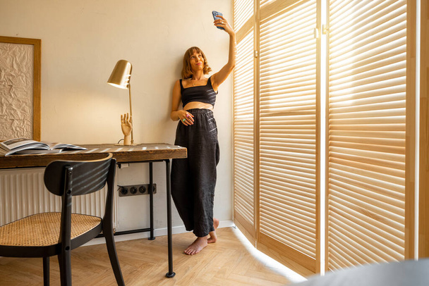 Młoda stylowa kobieta w czarnym ubraniu domowym rozmawiająca online lub robiąca zdjęcia na smartfonie stojąc w pobliżu żaluzji okiennych i miejsca pracy w domu. Pojęcie blogowania i stylu życia domowego - Zdjęcie, obraz