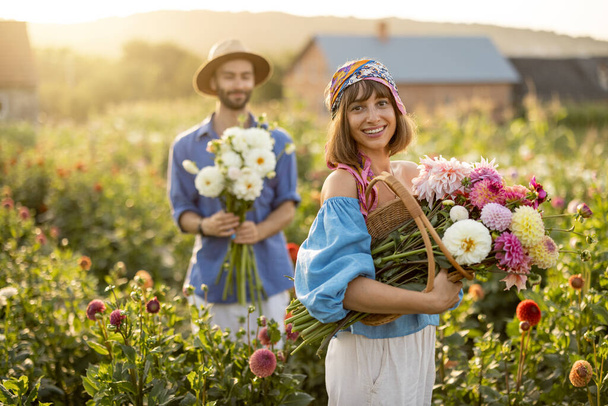 Portrait of cheerful man and woman stand as farmers with buckets full of freshly picked up dahlias while working at flower farm outdoors on sunset - Photo, Image