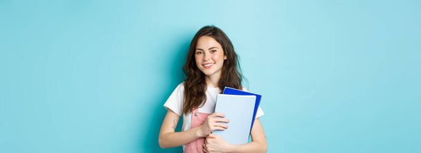 Cute smiling girl student holding notebooks and looking cheerful at camera, studying in college or university, standing over blue background. - Photo, image