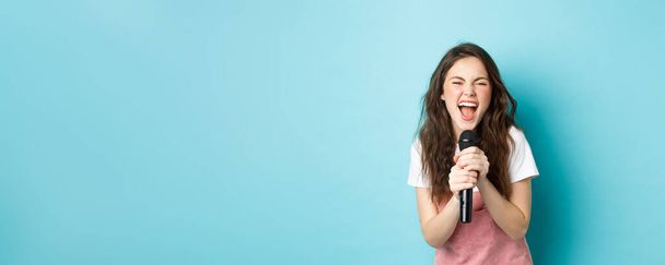 Aufgeregtes hübsches Mädchen, das Karaoke singt, Mikrofon in der Hand hält und glücklich lächelt, vor blauem Hintergrund stehend. - Foto, Bild
