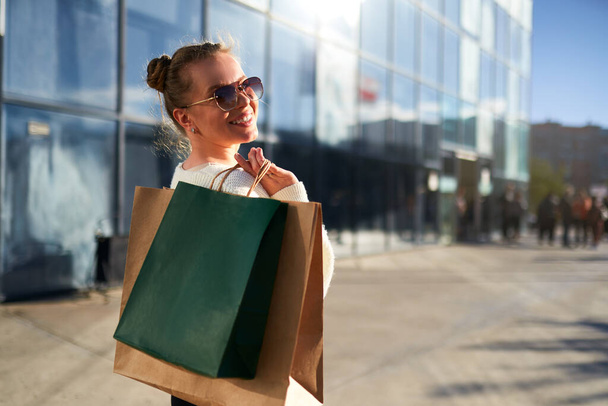 Happy woman with shopping bags walking in city from mall. Excited girl enjoys purchases on black friday sale. Female posing on camera with full paper bags. A consumerism concept. Back view. - 写真・画像