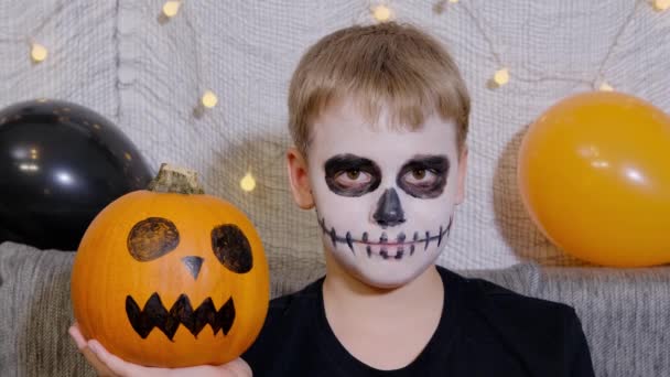 Portret van een tiener met make-up op zijn gezicht in de vorm van een skelet en met een pompoen in zijn handen. Het kind viert Halloween. - Video