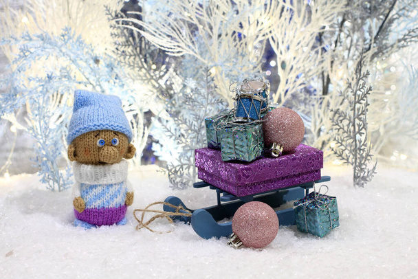人工雪の上にはニットのおもちゃのノームが立ち、白と青の人工木を背景にプレゼント付きのおもちゃのそりが彼の隣に立っている。閉鎖だ。ナイトスタジオ撮影, - 写真・画像