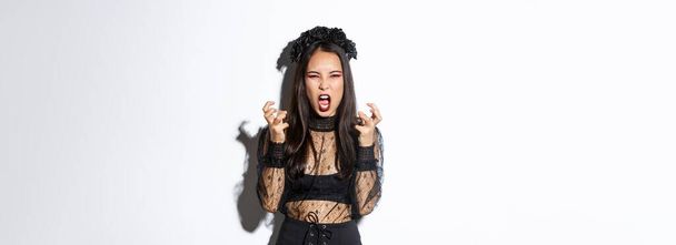 Image of mad woman in gothic lace dress and black wreath yelling angry and clenching fists, standing in halloween costume with furious expression, standing over white background. - Photo, Image